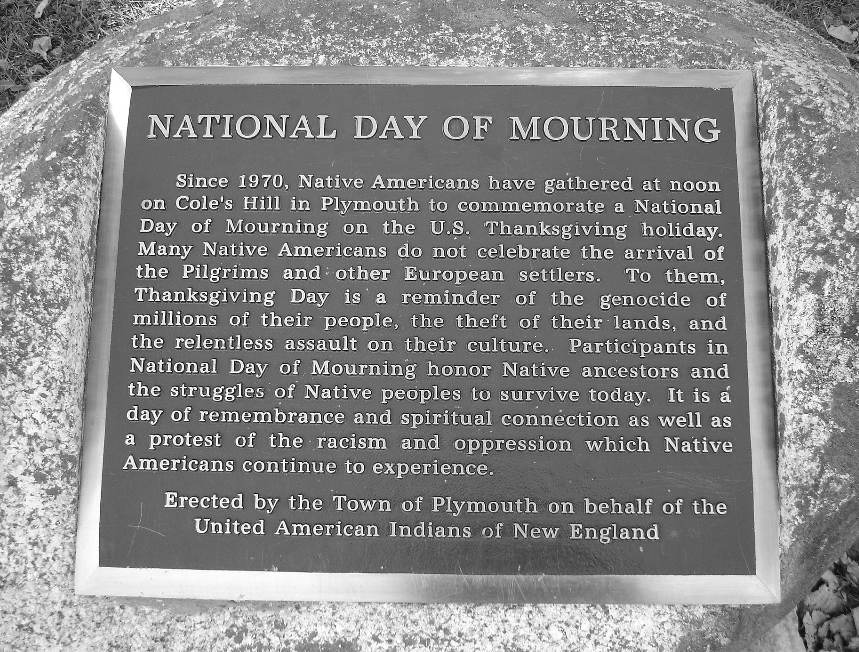 Image of a plaque that reads, 'National Day of Mourning: Since 1970, Native Americans have gathered at noon on Cole's Hill in Plymouth to commemorate a National Day of Mourning on the U.S. Thanksgiving holiday. Many Native Americans do not celebrate the arrival of the Pilgrims and other European settlers. To them, Thanksgiving Day is a reminder of the genocide of millions of their people, the theft of their lands, and the relentless assault on their culture. Participants in National Day of Mourning honor Native ancestors and the struggles of Native peoples to survive today. It is a day of rememberance and spiritual connection as well as a protest of the racism and oppression which Native Americans continue to experience. Erected by the Town of Plymouth on behalf of the United American Indians of New England.'